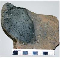 Brecha polimíctica com um fragmento de vidro de cor escura com  formato balístico. Esse fragmento exibe vesículas e amígdalas, sugerindo tratarse de material fundido pelo impacto durante sua trajetória balística, tendo se depositado junto com outros fragmentos para formar a brecha (Fonte: Crósta et al., 2013 - Registro SIGEP).

